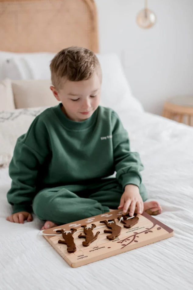 Child playing with a wooden puzzle on a bed, with a visible brand logo in the corner.